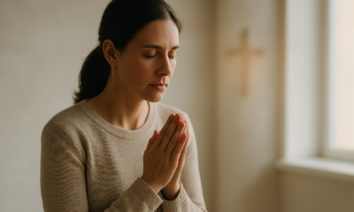 A woman quietly praying near a window with soft light and a blurred cross in the background