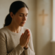 A woman quietly praying near a window with soft light and a blurred cross in the background