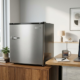A sleek modern mini refrigerator in a professional office environment, stainless steel finish, placed on a wooden cabinet next to a desk with laptop and coffee cup